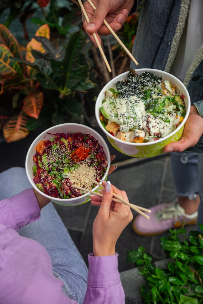 services-01 Two people holding colorful salad bowls with chopsticks, surrounded by greenery.